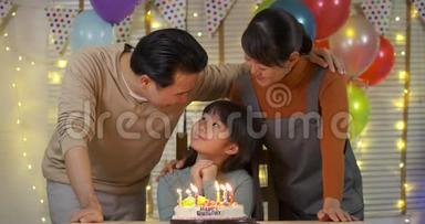 一个可爱的亚洲<strong>小女孩</strong>坐在桌子上吹着<strong>生日蛋糕</strong>上的蜡烛，而她的家人站在后面唱歌